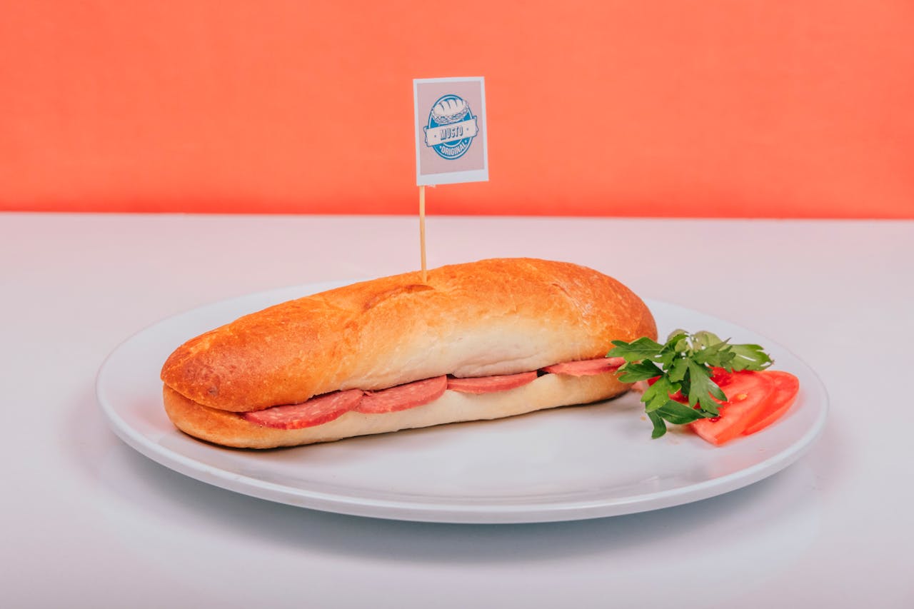 Tasty submarine sandwich with meat filling on white plate against orange background, garnished with tomato and parsley.